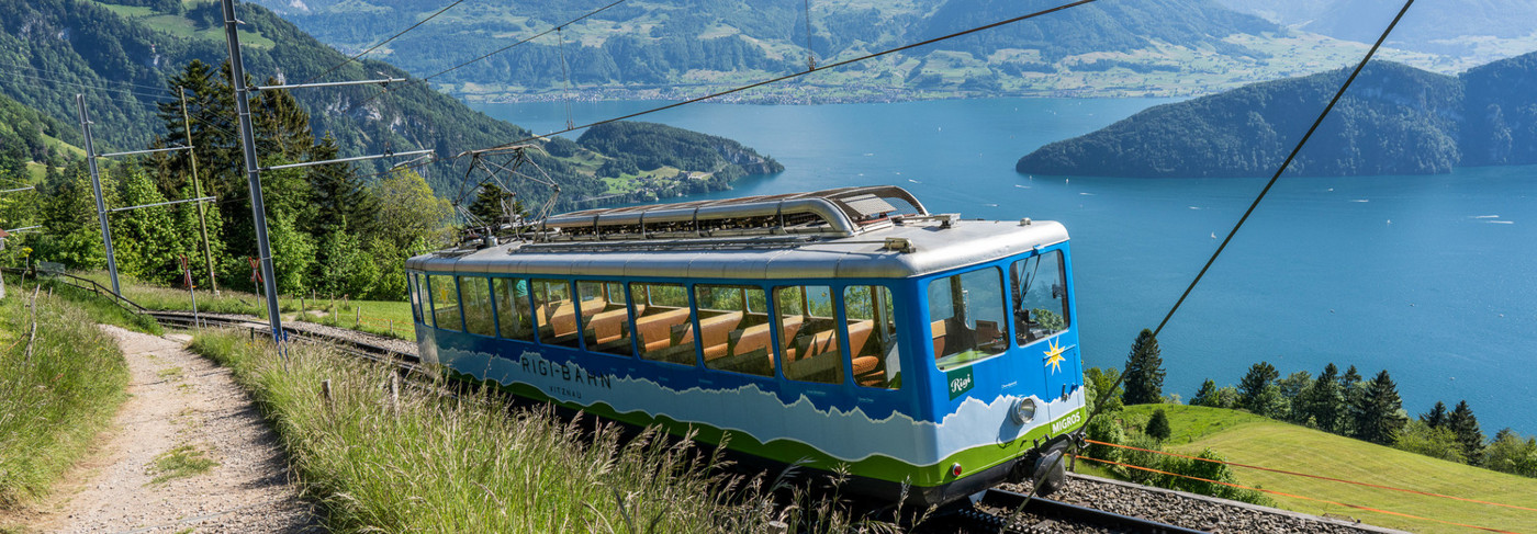 Eine blaue Rigi Bahn fährt die Rigi hoch mit dem Vierwaldstättersee im Hintergrund