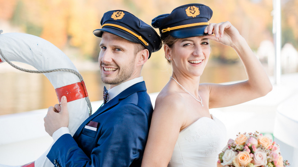 Schiffsmiete Hochzeit - Brautpaar Nahaufnahme vom Brautpaar. Beide haben einen Kapitänshut an und lächeln in die Kamera. Die Braut hält ihren Brautstrauss und der Bräutigam einen Rettungsring.