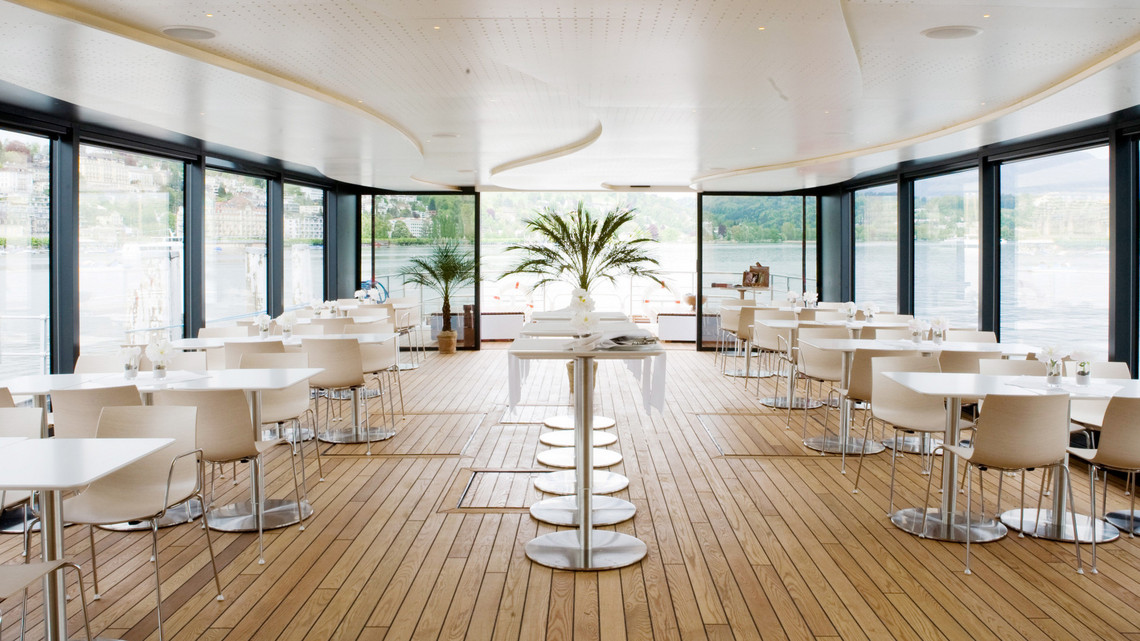 Salon du bateau à moteur Sapphire Sur le pont principal du bateau à moteur Saphir, il y a des tables et des chaises blanches à gauche et à droite des fenêtres panoramiques. Au milieu, il y a des tables de bar alignées.