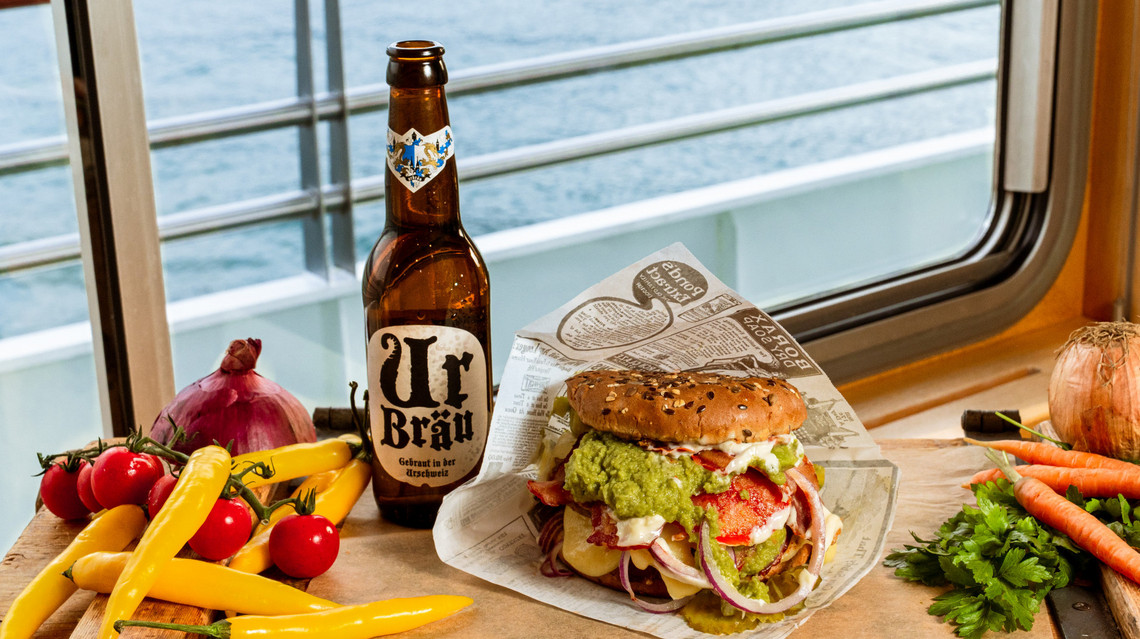 Auf dem Bild sieht man einen leckeren Burger mit Salat, Fleisch, Gemüse und Sauce. Neben dem Burger sieht man Ur Bräu Bier. Im Hintergrund sieht man Moos und Natur. 