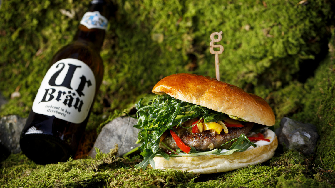 Burger Auf dem Bild sieht man einen leckeren Burger mit Salat, Fleisch, Gemüse und Sauce. Neben dem Burger sieht man Ur Bräu Bier. Im Hintergrund sieht man Moos und Natur.