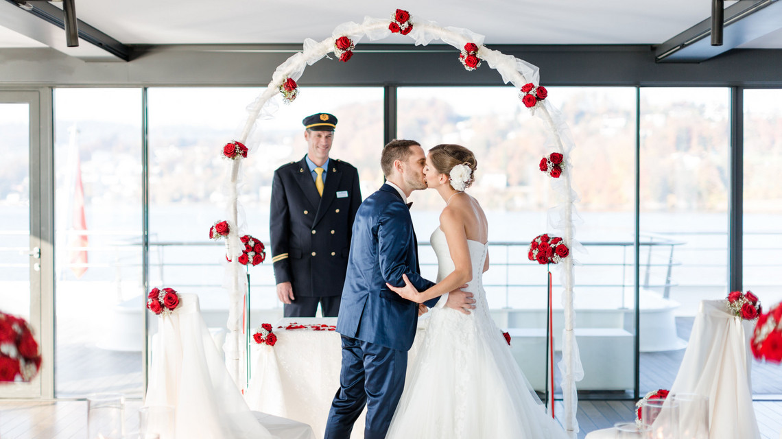 Schiffsmiete Hochzeit - Trauung Küssendes Brautpaar auf dem Schiff. Im Hintergrund sieht man den Kapitän lächlen sowie die Blumendekoration mit roten Rosen.